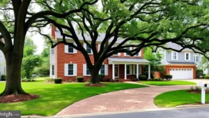 Charming colonial-style brick home with white shutters and mature oak trees in front yard, manicured lawn, brick driveway leading to garage, peaceful residential street in Virginia