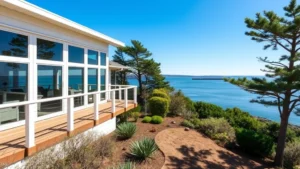 Modern coastal home with large windows overlooking Coos Bay water, white railings, landscaped yard with native plants and mature trees, bright sunny day with blue sky
